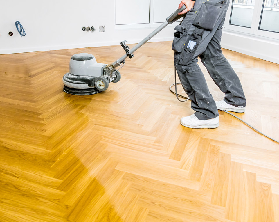 Polisher smoothing a wooden herringbone floor in a bright room.